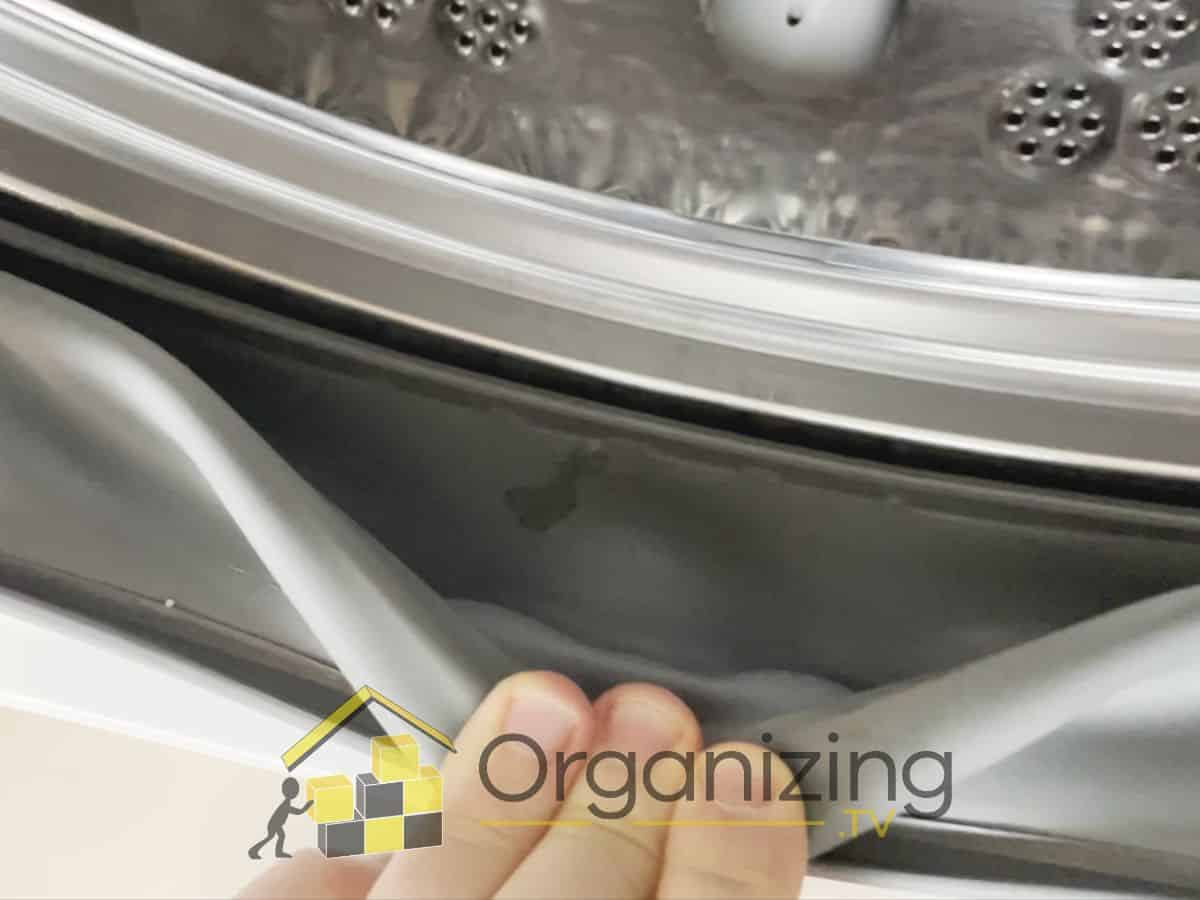 Hand pulling back the rubber gasket on a front-load washing machine to reveal hidden dirt and biofilm