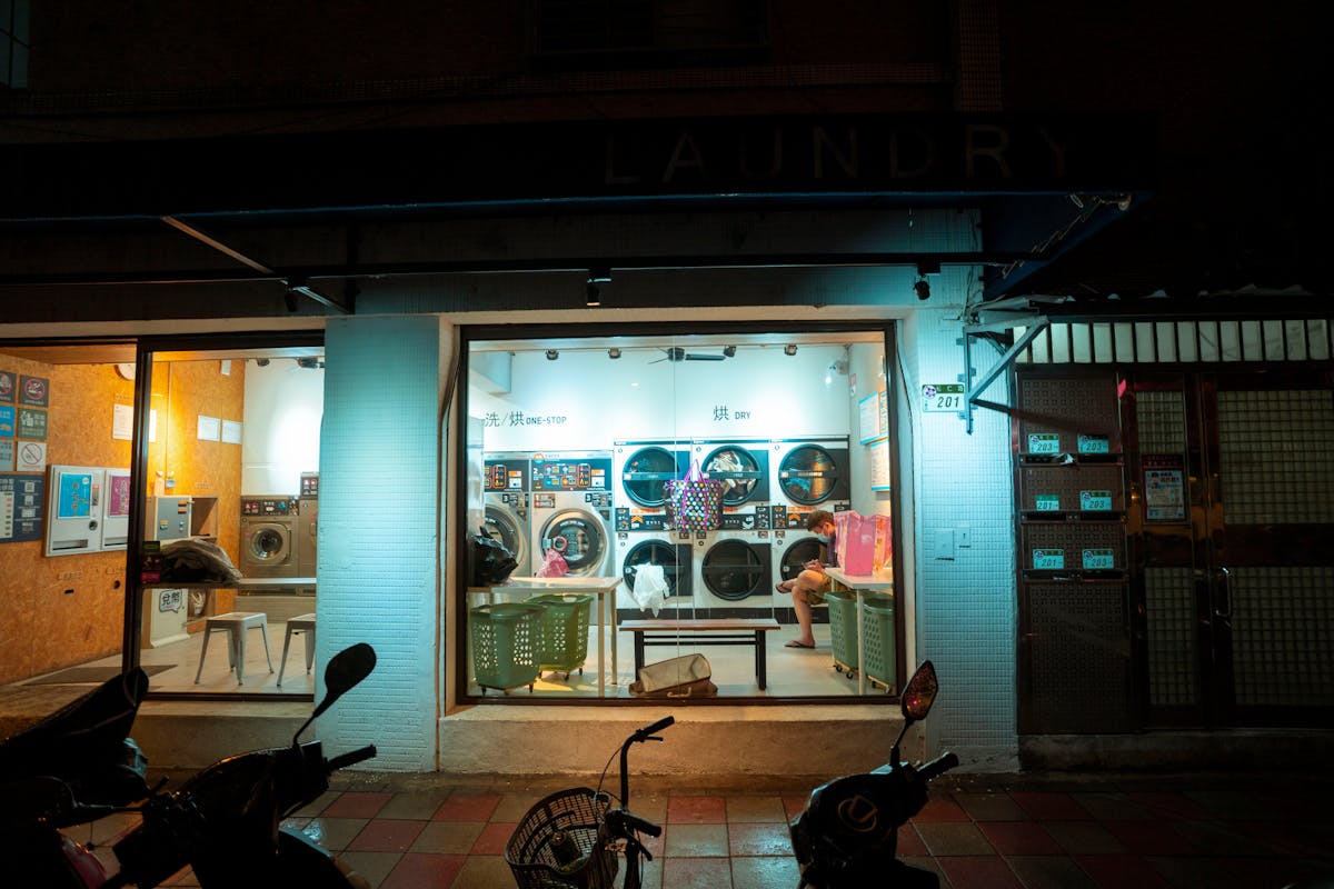 A brightly lit laundromat visible through its storefront window at night with washing machines and a person waiting inside