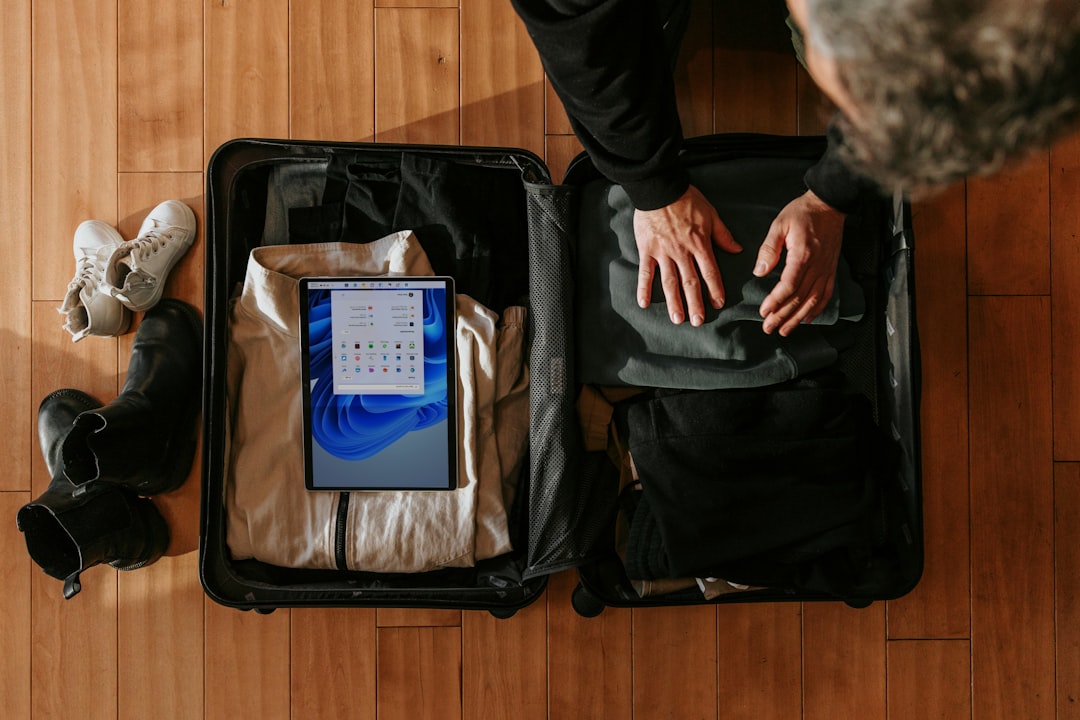 Person efficiently packing clothes and a tablet into an open carry-on suitcase on a wooden floor, with shoes placed nearby