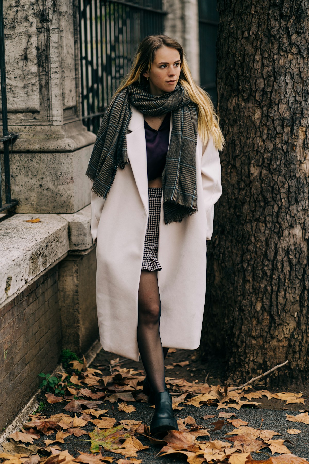 Woman wearing a cream coat over layers with a plaid scarf, walking through a European city with autumn leaves, showing layered dressing for variable weather