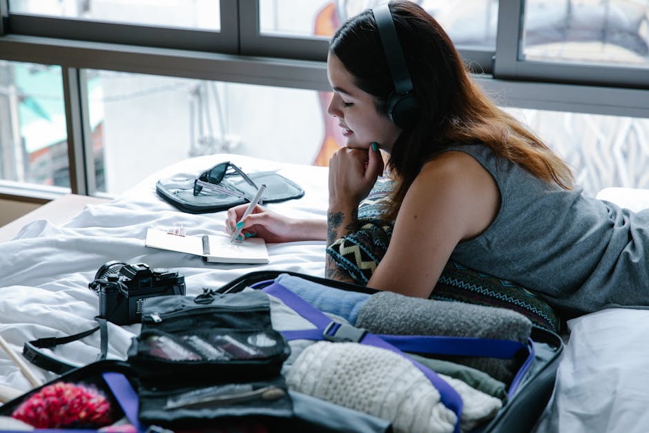 Woman lying on bed writing in a notebook next to an open suitcase reviewing what she packed for her trip
