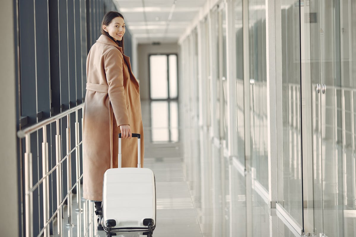 Stylish woman in a beige coat walking through an airport hallway with a white carry-on suitcase