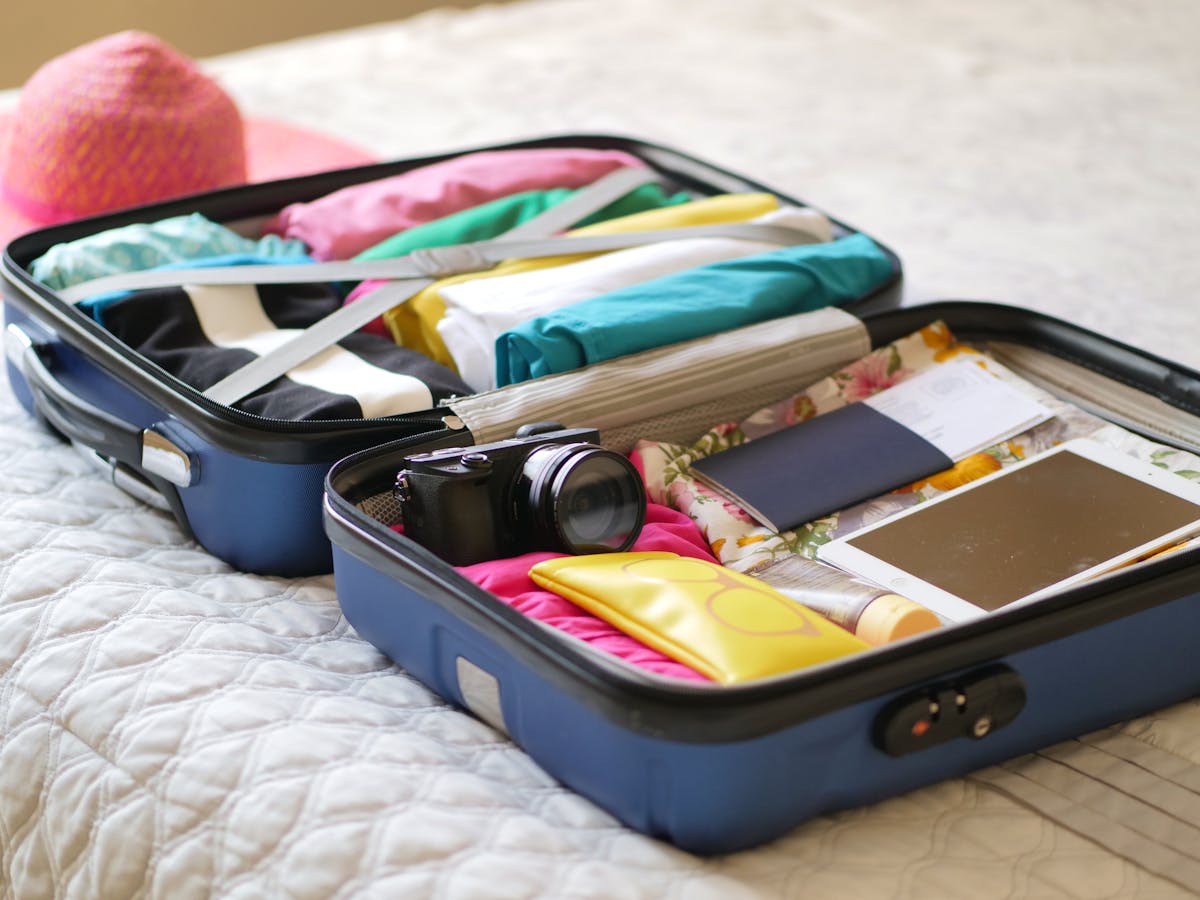 Open blue carry-on suitcase on a bed packed with folded colorful clothes, camera, tablet, and travel documents