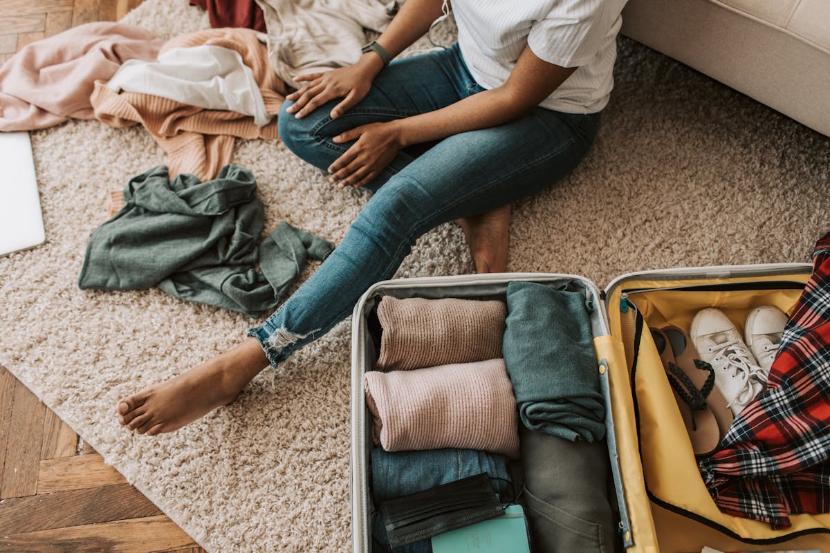 Woman sitting on floor packing rolled clothes and shoes into an open yellow carry-on suitcase