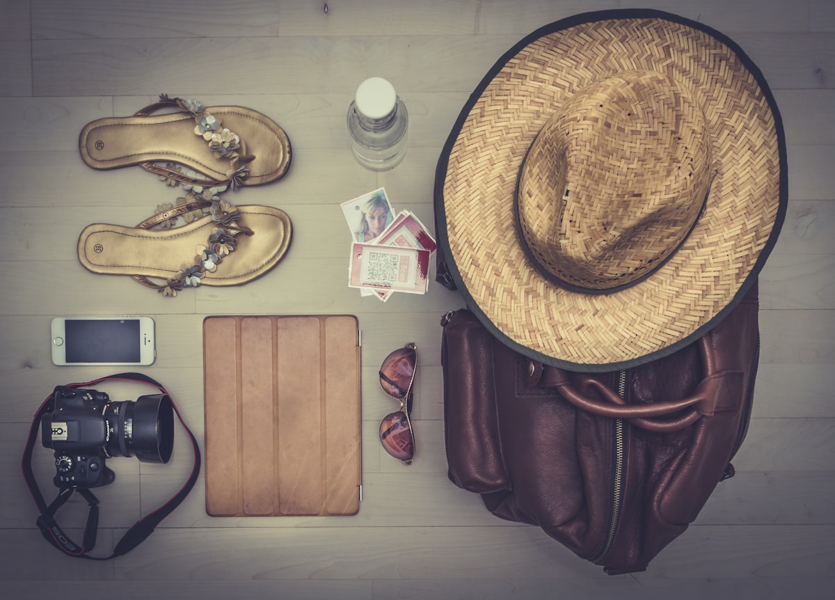 Flat lay of travel essentials including straw hat, sandals, camera, sunglasses, phone, and leather bag